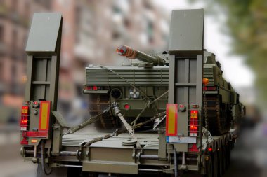 Büyük bir askeri aracın işlek bir şehir caddesinde bir topçu parçası taşıdığı görülüyor. Sahne, arka planda açık gökyüzünün altında binaların bulunduğu kentsel çevreyi tasvir ediyor..