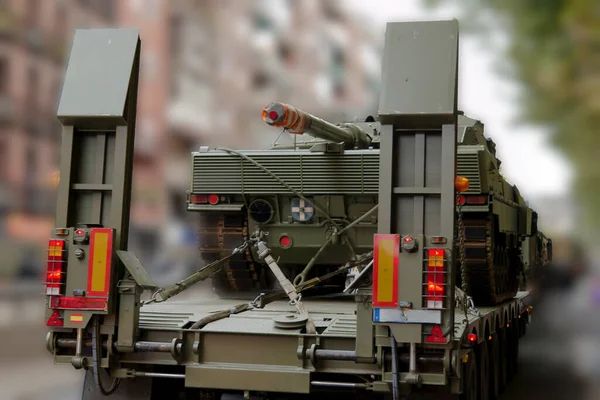 Büyük bir askeri aracın işlek bir şehir caddesinde bir topçu parçası taşıdığı görülüyor. Sahne, arka planda açık gökyüzünün altında binaların bulunduğu kentsel çevreyi tasvir ediyor..