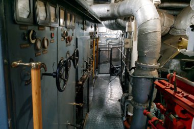 Tarihi bir geminin kontrol odasında, geniş boruların yanında çeşitli göstergeler ve kollar görülebilir. Mekan aydınlık, daha önceki zamanların karmaşık mühendislik detaylarını gösteriyor..