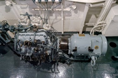 Bir geminin makine dairesinde jeneratörün yanında eski bir deniz motoru sergileniyor. Sahne karmaşık mekanik bileşenleri ve endüstriyel işçiliği yakalıyor..