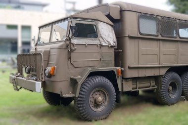 Engebeli bir tasarıma sahip askeri bir kamyon çimenli bir alana park edilmiş, sağlam tekerlekleri ve kamuflaj rengiyle sergileniyor. Modern binalar arka planda berrak bir gökyüzünün altında görülebilir..