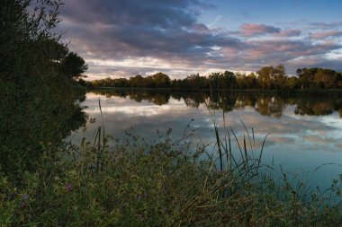 Sakin bir nehir manzarası alacakaranlığın güzelliğini yakalar. Ağaçlar akşam gökyüzünü yansıtırken, renkli bulutlar gökyüzünde sürükleniyor..
