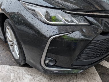 Siyah bir araba, bir şehrin yaya geçidinin yanına park edilmiş olarak yüzeyinde yağmur damlacıkları gösterir. Sahne şehir yaşamını ve yağmurlu havayı yakalıyor.