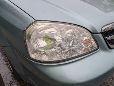 Araba farı yakından bakınca, mavi bir araçta karmaşık mercek tasarımları ve su damlacıkları görülüyor. Sahne, yansımaları ve berraklığı vurgulayan yağmurlu bir atmosfer yakalıyor..