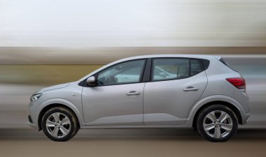 Parlak gümüş renkli bir araba, camlarında yağmur damlalarıyla yol boyunca hızla ilerliyor. Arka plan bulanık, hızlı hareket olduğunu gösteriyor. Su damlacıkları ışığa yakalanır ve sahneye dinamik bir hava katar..