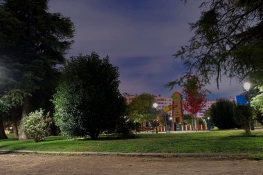 Bir şehir parkı, alacakaranlıkta ağaçlarla çevrili parlak oyun yapılarıyla dolu canlı bir oyun alanı gösteriyor. Sokak lambaları usulca parlıyor, hoş bir atmosfer yaratıyor..