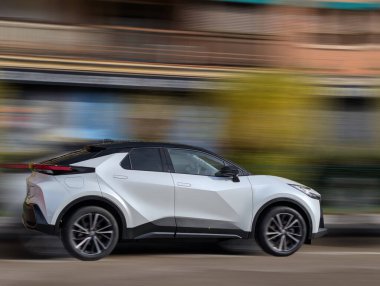 Modern beyaz bir çapraz araba şehir caddesi boyunca hızlanır. Parlak binalar açık mavi gökyüzünün altında canlı bir zemin oluşturarak şehir yaşamının ve hızının özünü yakalıyor..