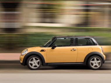 Küçük sarı bir araba şehir caddesinde hızla ilerliyor, hareket bulanıklığı gösteriyor. Sahne, arka planda yeşillik olan canlı şehir yaşamını yakalıyor..