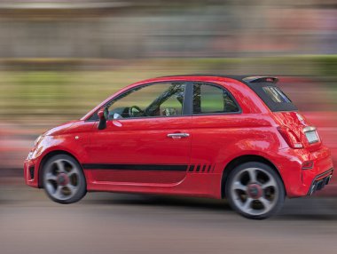 Canlı kırmızı kompakt bir araba şehir yolunda hızlanarak spor tasarımını sergiliyor. Bulanık yeşillik gün ışığında bir hareket hissi yaratır..