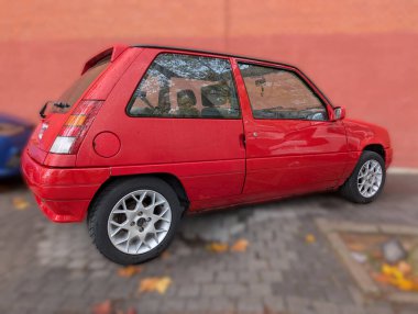 Canlı kırmızı bir kompakt araba renkli bir duvarın yanında park edilmiştir. Mevsimsel değişimi vurgulamak için sonbahar yaprakları asfalta saçılmıştır. Sahne, sakin bir şehir anını yakalıyor..