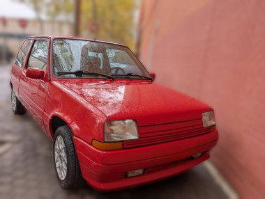 Yağmurlu bir günde parlak kırmızı bir araba renkli bir duvarın yanında park edilir. Gri gökyüzü tepemizde belirdiğinde yağmur damlaları yüzeyinde parlar..