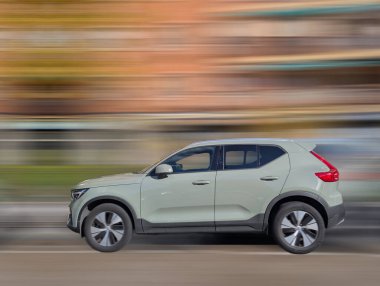 Gösterişli bir araba hareketli bir şehir caddesinde hızla ilerliyor, arka planda bulanık binalar yüksek hızda olduğunu gösteriyor. Sahne, gündüz vakti bir heyecan ve enerji hissi yakalıyor..