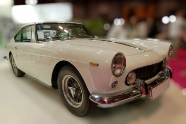 Klasik bir beyaz spor araba bir otomobil fuarında sergileniyor, gösterişli çizgilerini ve klasik ayrıntılarını vurguluyor, heveslilerle dolu hareketli bir sergi salonu arasında.