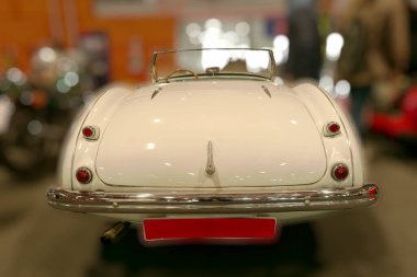 Klasik beyaz üstü açık araba kapalı bir otomobil fuarında sergileniyor. Gösterişli tasarım ve cilalı vücut klasik otomobillerin çekiciliğini yansıtıyor, araba tutkunlarını ve koleksiyoncuları cezbediyor..