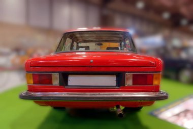 Bir otomobil fuarı sırasında bir sergi salonunda kırmızı bir araba sergileniyor. Odak noktası arabanın arkası, tasarımı ve rengi. Ayarlar yeşil zemin ve çeşitli araçları içeriyor.