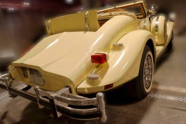 Üstü açık bir araba garajda duruyor. Arabanın parlak sarı bir vücudu, krom detayları ve klasik tasarım unsurları var. Hareket etmeden park halinde, özelliklerini vurguluyor..