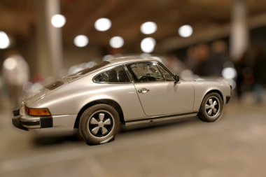 Arka planda bulanık kalabalık olan bir mekanda gümüş renkli bir spor araba sergileniyor. Işıklandırma modern alanda arabanın hatlarını ve parlak çizgilerini vurguluyor..