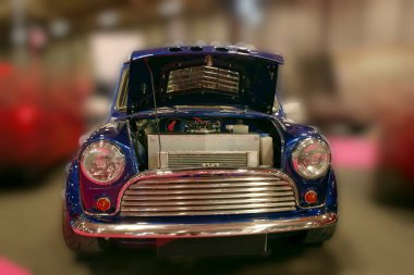 Şehirdeki bir otomobil fuarında kaputu açık duran klasik bir mini araba. Ziyaretçiler olayın canlı atmosferinin tadını çıkarırken motoru gözlemliyorlar.