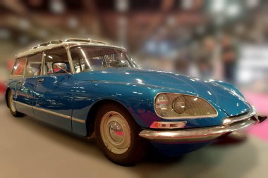 Mavi bir klasik araba, insanların klasik araçlara hayran olduğu bir otomotiv etkinliğinde park edilir. Etkinlikte çeşitli modeller yer alıyor ve arabalarla ilgilenen çok sayıda ziyaretçi çekiyor..