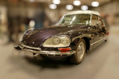 Klasik bir araba, retro temalı bir mekanda sergileniyor. Araç zarif kıvrımlar ve parlak detaylar içeriyor. İnsanlar canlı bir araba şovu sırasında tasarımı beğenerek etrafta dolanıyorlar..