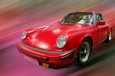 Klasik kırmızı bir araba bir otomobil fuarında sergilenir. Arka plan bulanık görünürken araba odak noktasında duruyor. Bu da canlı bir atmosfer olduğunu gösteriyor. Ziyaretçiler araca hayran hayran hayran kalarak geçiyorlar..