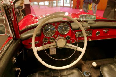 Kırmızı klasik bir arabanın içi klasik bir direksiyon ve torpido gözüdür. Detaylar, bir araba fuarında dikkat çeken tasarım ve işçiliği vurguluyor..