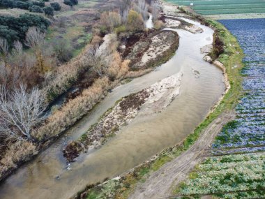 Ağaçlar ve tarımsal alanlarla dolu bir arazide bir nehir akıyor. Su kıyılar boyunca yavaşça hareket eder. Sahne, doğa ve çiftçilik faaliyetlerinin karışımını gösteriyor..