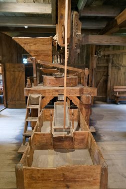 Tahta tahıl değirmeni ekipmanları kırsal bir binada duruyor ve geçmişin işçiliğini sergiliyor. Yapı tahıl işlemenin tarihsel sürecini vurguluyor.