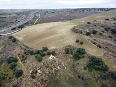 Hava manzarası, bulutlu bir gökyüzünün altındaki tarım arazilerini ve yuvarlanan tepeleri gösteriyor. Manzaranın içinden bir otoyol geçiyor. Ağaçlar tarlaları dolduruyor, kırsal yaşamı sergiliyor..