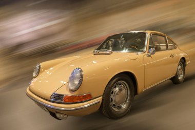 Açık kahverengi klasik bir araba bulanık çevrelerle dolu bir mekanda yarışıyor. Etkinlikte tasarım ve tarihlerini vurgulayarak çeşitli vintage araçlar sergilenecek.