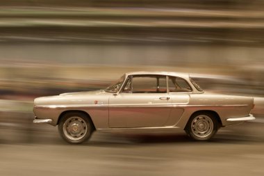 Öğleden sonra işlek bir şehir caddesi boyunca klasik bir gümüş araba hız yapar. Arka plan bulanık, hareket gösteriyor. Yakınlarda insanlar ve binalar görülebilir..