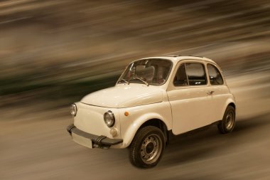 Küçük beyaz klasik bir araba şehir ortamında yol boyunca hızlı hareket eder. Arka planda hareket bulanıklığı görülüyor. Olay yerindeki hız ve aktiviteyi gösteriyor..