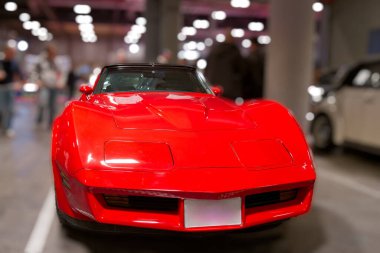 Kırmızı bir spor araba bir otomobil fuarında duruyor. İnsanlar etrafta dolanıp arabaya bakıyorlar. Olay kapalı alanda geniş bir alanda gerçekleşir. Işık yukarıdan gelir..
