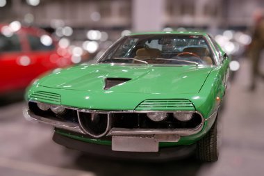 Yeşil klasik bir araba bir otomotiv etkinliği sırasında büyük bir salonun içinde sergilenir. Arka planda başka arabalar da görülebilir ve ışıklandırma parlak..