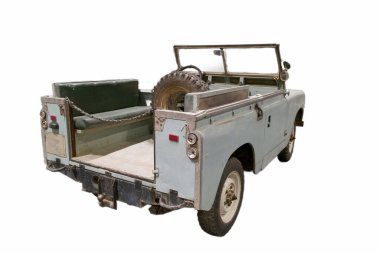 Klasik bir off road aracı garajda duruyor. Arka tarafta iki koltuk ve bir yedek lastik var. Üstü açık ve güneş ışığı alanı dolduruyor. Aracın tasarımını vurguluyor..