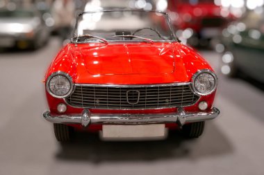 Büyük bir sergi salonunda klasik kırmızı üstü açık bir araba sergileniyor. İnsanlar vitrinde yürüyor, araç gösterisi sırasında klasik araçlara hayran bakıyorlar..