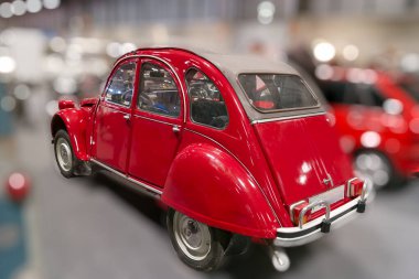 Klasik kırmızı bir araba bir otomobil fuarında sergilenir. Aracın kavisli bir tasarımı var ve iç kısmı görülebilir. Arka planda diğer arabalar bulanık..