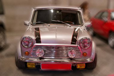 Klasik bir mini araba, yüzeyinden yansıyan parlak ışıklarla dolu bir garajda park halinde. Arka planda diğer arabalar görünürken araba krom detaylar ve canlı renkler gösteriyor..