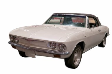 Beyaz, üstü açık bir araba kapalı bir mekanda bir otomotiv etkinliğinde sergileniyor. Arabanın siyah bir üstü ve parlak tekerlekleri var. Birçok insan farklı araçlara bakıyor..