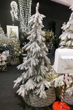 İki yapay karlı Noel ağacı büyük sepetlerde duruyor. Dekorasyonlu bir iç mekandaki şenlikli gösterinin bir parçası. Işıklar arka planda usulca parlıyor, tatil ruhunu güçlendiriyor..