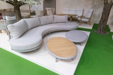 Modern kapalı bir alanda dairesel bir kanepe bulunur. Açık gri kumaş kanepeyi kaplıyor ve yakınlarda yuvarlak bir ahşap masa oturuyor. Yerde yeşil halı var. Başka mobilyalar da görünür durumda..