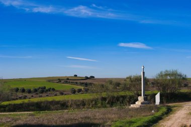 Taştan bir anıt kırsal bir alanda toprak bir yolun kesişiminde duruyor. Uzakta tepeler ve açık mavi gökyüzü ile, her iki tarafta yeşil tarlalar uzanır.