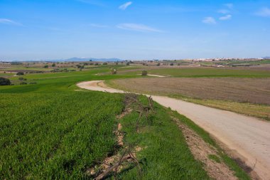 Toprak bir yol, açık bir gökyüzünün altındaki yeşil çimlerin ve kahverengi toprağın arasından esiyor. Tepeler arka planda görülebilir ve uzakta küçük bir kasaba vardır. Güneş ışıldıyor.