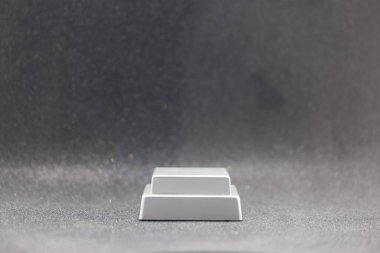 Beyaz dikdörtgen bir cisim gri bir yüzeyde bulunur. Arka planda dokulu bir görünüş ve hafif bir parıltı var. Işık, nesnenin temiz çizgilerini vurguluyor ve keskin bir kontrast oluşturuyor..