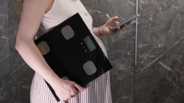 Hamile bir kız elinde elektronik bir terazi tutuyor ve ağırlığını ölçmek istiyor.
