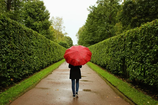En ung flicka promenader längs de gröna gränderna från buskarna i regnet med ett rött paraply — Stockfoto