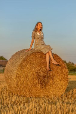 Mavi elbiseli genç kadın gün batımında samanlıklarla arka planda yakın çekim yapıyor..