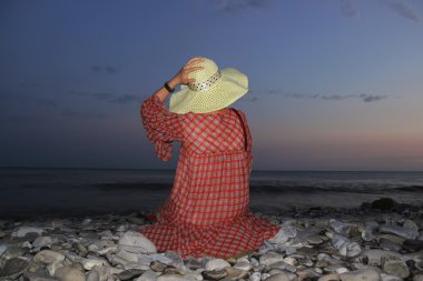 Genç güzel kız deniz kıyısında günbatımı ve şapkasını eliyle tutarak oturur