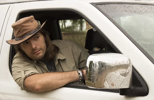 Handsome young man with long hair in brown cowboy hat driving white 4x4 car after rain. Safari. Country style. romance.