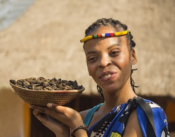 Sudáfrica, Gauteng, Lesedi Cultural Village (centro único de la cultura africana) 04 de julio de 2015. Joven sonriente nación bantú sirviendo orugas comestibles para la cena. Muchacha mostrando cesta de orugas en su palma. Deliciosa comida inusual . — Foto de Stock